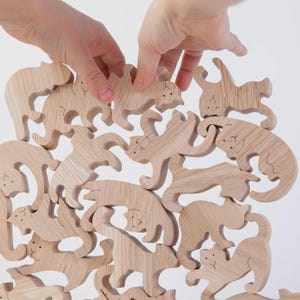 Könnte beinhalten: Holzpuzzleteile in Katzenform in verschiedenen Posen, mit Händen, die ein Teil platzieren. Das hellfarbene Holz hat sichtbare Maserungen und einfache Gesichtszüge, die in jede Katze geschnitzt sind. Ein spielerisches, lehrreiches Spielzeug.