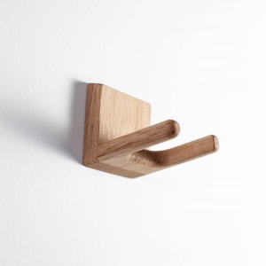 Puede incluir: Un gancho de pared de madera con una base cuadrada y dos brazos curvos. El gancho es de madera clara y est&aacute; montado en una pared blanca. El dise&ntilde;o es minimalista y moderno, ideal para colgar objetos.