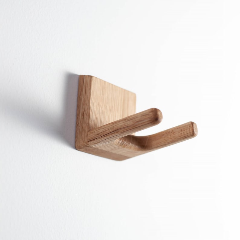 Puede incluir: Un gancho de pared de madera con una base cuadrada y dos brazos curvos. El gancho es de madera clara y est&aacute; montado en una pared blanca. El dise&ntilde;o es minimalista y moderno, ideal para colgar objetos.