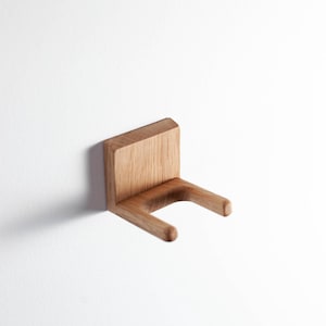 Puede incluir: Soporte de pared de madera clara con dise&ntilde;o minimalista. Cuenta con una parte trasera rectangular y dos brazos redondeados. Montado en una pared blanca, este soporte de madera presenta un dise&ntilde;o sencillo y moderno, ideal para diversos usos.
