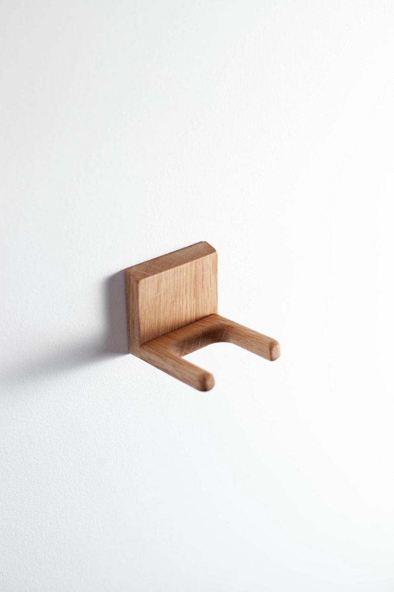 Puede incluir: Soporte de pared de madera clara con dise&ntilde;o minimalista. Cuenta con una parte trasera rectangular y dos brazos redondeados. Montado en una pared blanca, este soporte de madera presenta un dise&ntilde;o sencillo y moderno, ideal para diversos usos.