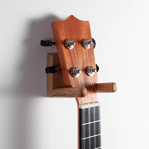 Könnte beinhalten: Eine Wandhalterung aus Holz für eine Ukulele, die Kopfplatte und einen Teil des Halses zeigt. Die Halterung ist aus hellem Holz gefertigt, das sich von der rotbraunen Kopfplatte abhebt. Die Saiten und Stimmwirbel sind sichtbar.