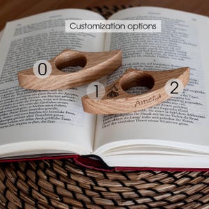 K&ouml;nnte beinhalten: Zwei gebogene Buchst&uuml;tzen aus Holz, die auf einem offenen Buch liegen. Eine hat ein Blumendesign, die andere den Namen "Amelia" eingraviert. Der Text "Customization options" steht oben.
