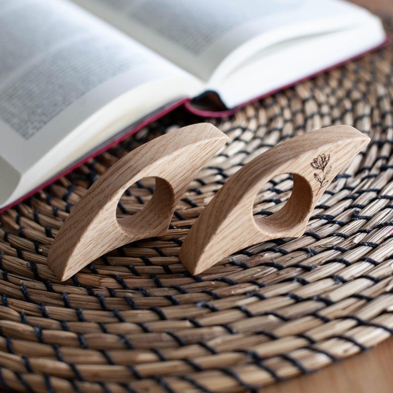 K&ouml;nnte beinhalten: Zwei Buchhalter aus hellem Holz. Einer hat ein kleines Blumendesign. Die Halter sind so konzipiert, dass sie &uuml;ber einen Finger geschoben werden, um ein Buch offen zu halten. Das Buch liegt auf einer gewebten Oberfl&auml;che.
