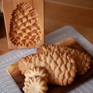 Könnte beinhalten: Eine hölzerne Keksform und gebackene Kekse in Form von Ananas und Sternen. Die Form ist rechteckig mit einem geschnitzten Ananas-Design. Die Kekse sind goldbraun und liegen auf einem Holzbrett.