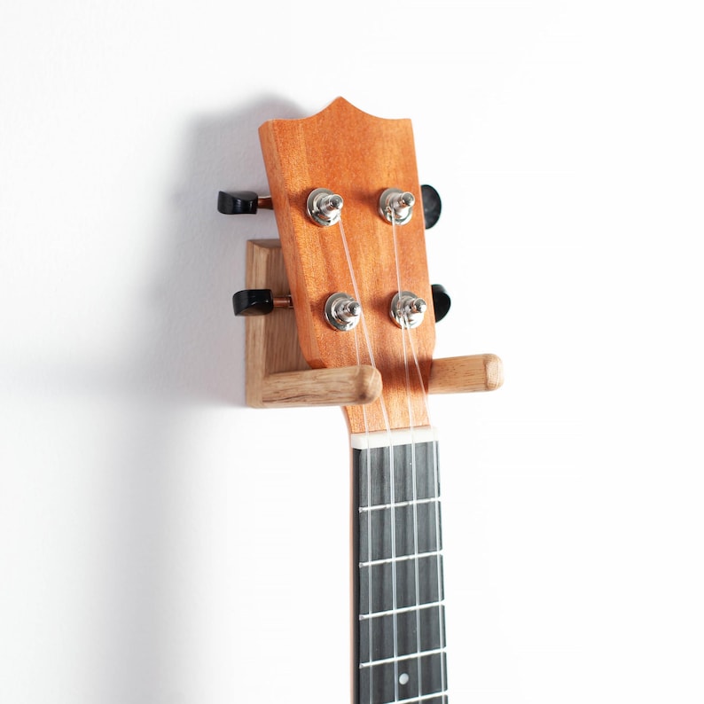 Puede incluir: Un soporte de pared de madera para ukelele, que muestra el clavijero del instrumento y una parte del diapas&oacute;n. El soporte es de madera clara, con el clavijero del ukelele en un tono marr&oacute;n rojizo c&aacute;lido. Se ven las cuerdas y las clavijas.