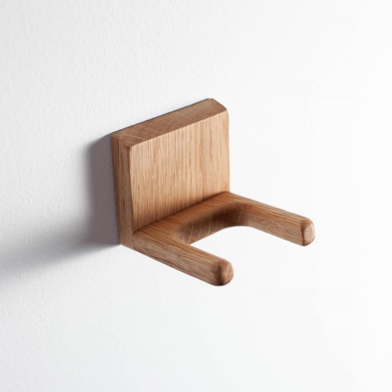 Puede incluir: Un soporte de pared de madera con acabado natural. El soporte presenta una parte trasera rectangular y dos brazos curvos que se extienden hacia afuera. El dise&ntilde;o es minimalista y moderno, adecuado para varios art&iacute;culos.