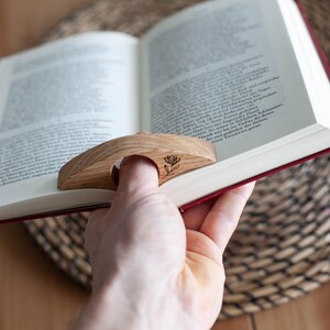 K&ouml;nnte beinhalten: Ein h&ouml;lzerner Daumen-Seitenhalter mit floralem Design liegt auf einem offenen Buch. Der Seitenhalter ist aus hellem Holz gefertigt und hat eine gebogene Form mit einem Loch f&uuml;r den Daumen. Das Buch hat rote Kanten.