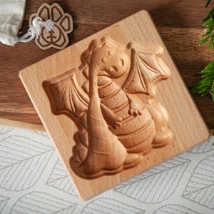 Peut inclure: Un moule à biscuits en bois en forme de dragon. Le moule est sculpté avec un motif de dragon détaillé, avec des ailes et un long cou. Le moule est sur une surface en bois, avec une petite étiquette en bois en forme de patte et une branche de conifère.