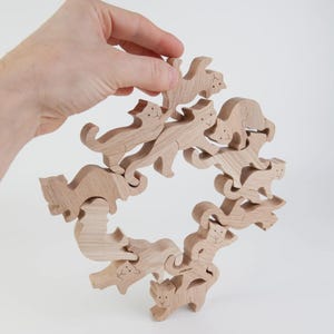 Op de afbeelding: Een houten puzzel met meerdere kattenfiguren die in elkaar grijpen om een ruitvorm te vormen. De katten zijn lichtbruin met geëtste gezichtskenmerken. De puzzel wordt door een hand tegen een witte achtergrond gehouden.