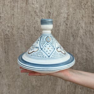 Puede incluir: Una olla de tagine de cer&aacute;mica blanca y azul con tapa c&oacute;nica. La tapa y la base presentan intrincados patrones geom&eacute;tricos y detalles dorados. El tagine se sostiene en la mano de una persona contra un fondo texturizado.