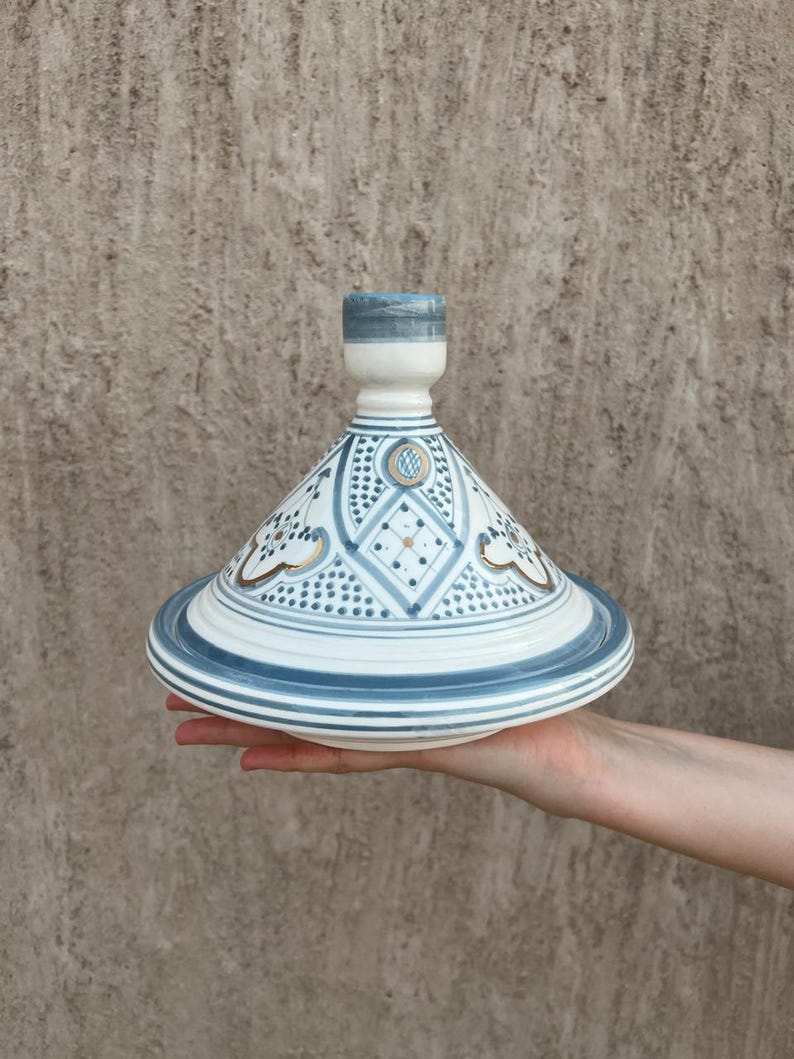 Puede incluir: Una olla de tagine de cer&aacute;mica blanca y azul con tapa c&oacute;nica. La tapa y la base presentan intrincados patrones geom&eacute;tricos y detalles dorados. El tagine se sostiene en la mano de una persona contra un fondo texturizado.