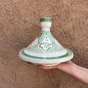 Puede incluir: Una olla de tagine de cer&aacute;mica blanca y verde con tapa c&oacute;nica y base ancha. La tapa presenta un patr&oacute;n geom&eacute;trico con detalles en verde y dorado. La olla se sostiene en la mano de una persona sobre un fondo marr&oacute;n texturizado.
