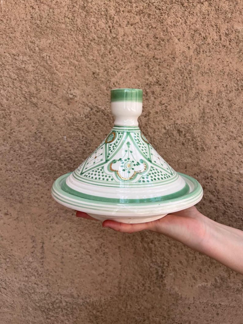 Puede incluir: Una olla de tagine de cer&aacute;mica blanca y verde con tapa c&oacute;nica y base ancha. La tapa presenta un patr&oacute;n geom&eacute;trico con detalles en verde y dorado. La olla se sostiene en la mano de una persona sobre un fondo marr&oacute;n texturizado.