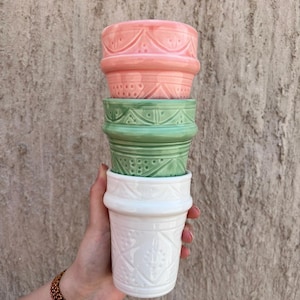 May include: A stack of ceramic plant pots in pink, green, and white. Each pot has a different embossed pattern. The pots are held in a hand, highlighting the decorative design and colour variations. The pots are suitable for indoor or outdoor use.