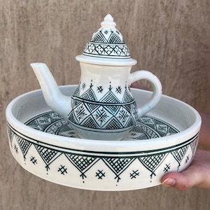 Peut inclure: Théière en céramique blanche avec un couvercle assorti et un plateau de service rond. La théière et le plateau sont décorés d'un motif géométrique vert foncé. La théière a une anse et un bec. Le plateau est tenu par une personne.