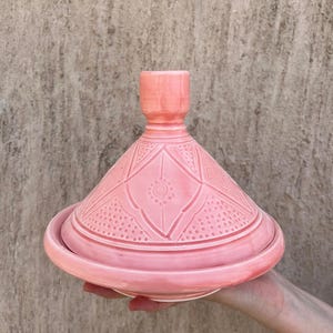 Puede incluir: Una olla de tagine de cer&aacute;mica rosa con tapa c&oacute;nica y base redonda. La tapa presenta un patr&oacute;n geom&eacute;trico. La olla se sostiene sobre un fondo neutro. Esta cocina tradicional se utiliza para guisos y tagines a fuego lento.