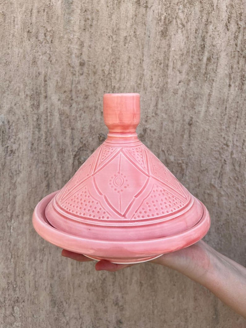 Puede incluir: Una olla de tagine de cer&aacute;mica rosa con tapa c&oacute;nica y base redonda. La tapa presenta un patr&oacute;n geom&eacute;trico. La olla se sostiene sobre un fondo neutro. Esta cocina tradicional se utiliza para guisos y tagines a fuego lento.