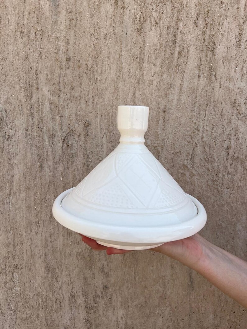Puede incluir: Tajine de cer&aacute;mica blanca con tapa c&oacute;nica y base ancha y redonda. La tapa presenta un patr&oacute;n geom&eacute;trico. El tajine se sostiene en la mano de una persona sobre un fondo neutro. Un recipiente de cocina tradicional.