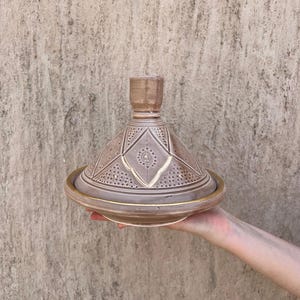 Puede incluir: Una olla de tagine de cer&aacute;mica marr&oacute;n con tapa c&oacute;nica y base ancha. La tapa presenta patrones geom&eacute;tricos y un peque&ntilde;o asa. El borde de la base tiene un acento dorado. El tagine se sostiene frente a una pared texturizada.