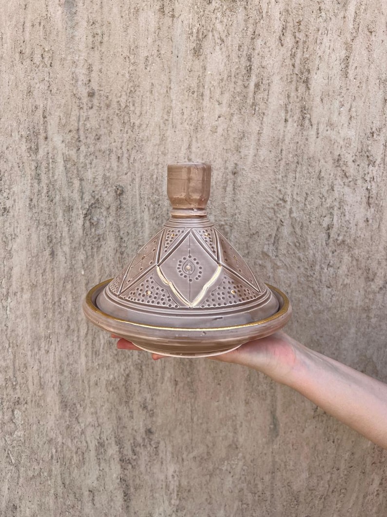 Puede incluir: Una olla de tagine de cer&aacute;mica marr&oacute;n con tapa c&oacute;nica y base ancha. La tapa presenta patrones geom&eacute;tricos y un peque&ntilde;o asa. El borde de la base tiene un acento dorado. El tagine se sostiene frente a una pared texturizada.
