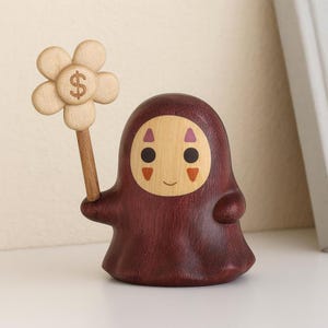 Op de afbeelding: Houten beeldje van een personage met een bordeauxrode body en een licht houten gezicht. Het personage houdt een bloem vast met een dollarteken in het midden. Het beeldje heeft een lachende uitdrukking en is waarschijnlijk een decoratief item.