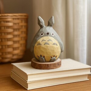 May include: A hand-carved wooden Totoro figurine, painted in gray and yellow, sits on a small wooden base atop two books. The figurine has large eyes and a detailed belly. The background includes a woven basket and a curtain.