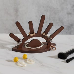 Puede incluir: Un organizador de joyas de madera marrón oscuro con forma de ojo, que sostiene anillos y una pulsera de perlas. También hay pendientes amarillos y brochas de maquillaje. El organizador tiene múltiples ranuras para anillos.