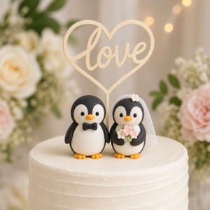 Puede incluir: Un adorno para pastel con dos figuras de pingüinos sobre un pastel blanco. Un pingüino lleva una pajarita negra, el otro lleva un velo blanco y sostiene un pequeño ramo de flores. Encima hay un letrero en forma de corazón con la palabra love.