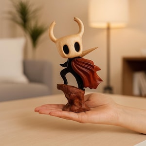 May include: A wooden figurine of a character with a light tan head, large black eyes, and curved horns. The figure wears a black body and a red cape, standing on a brown base. The figurine is held in a person's hand.