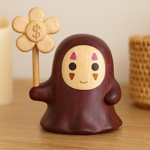 Op de afbeelding: Houten beeldje van een personage met een ronde kop en een donkerbruin lichaam. Het gezicht is lichtgekleurd met roze accenten en zwarte ogen. Het personage houdt een houten bloem vast met een dollarteken in het midden.