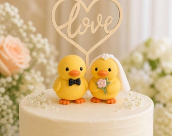 May include: A cake topper featuring two yellow duck figurines standing on a white cake. One duck wears a black bow tie, and the other wears a white veil and holds a small pink flower. Above them is a wooden heart cutout with the word love written.