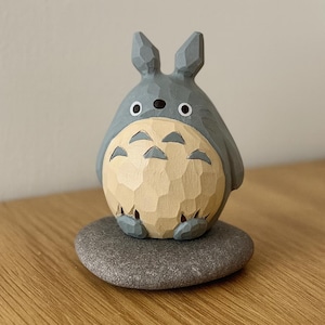 May include: A carved wooden figurine of Totoro, a character from the animated film My Neighbor Totoro. The figure is gray with a cream-colored belly and sits on a gray stone base. The figurine has large eyes and pointed ears.