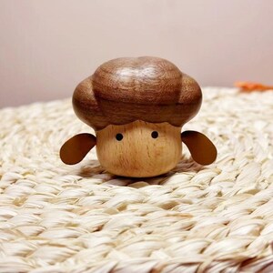 May include: A wooden mushroom figurine with a brown cap and a light brown body. The figurine has two small, round black eyes and two small, brown ear-like appendages. The mushroom is sitting on a woven, light-colored surface.