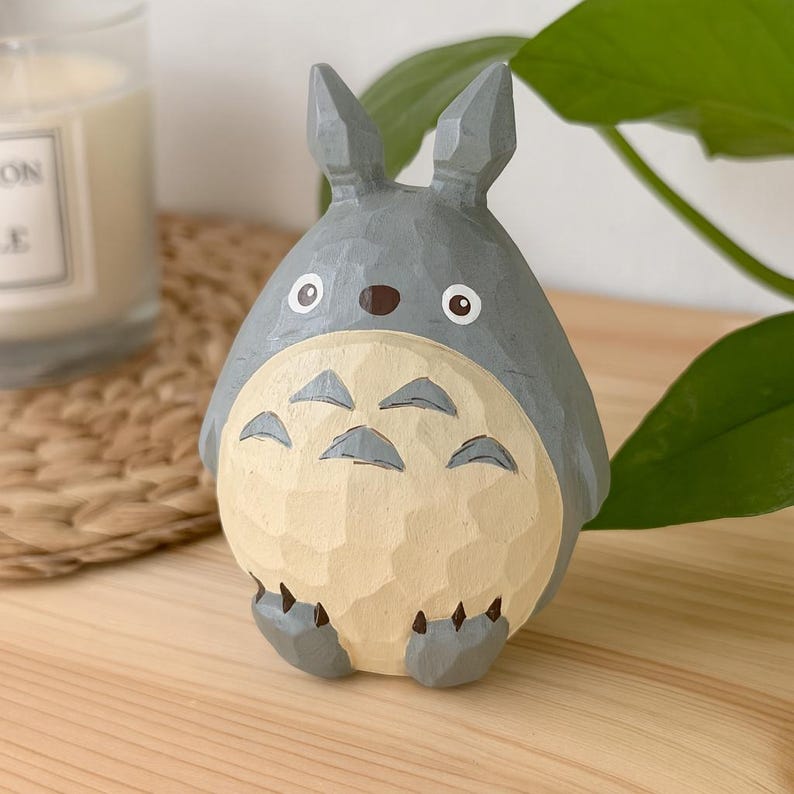 May include: A gray and cream-colored wooden Totoro figurine. The figurine has large ears, a round body, and a textured belly. It has black claws and a brown nose. The figurine is sitting on a wooden surface.
