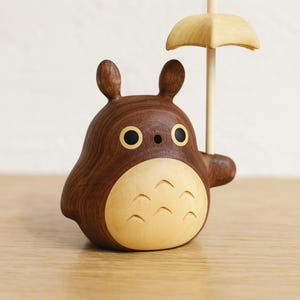 May include: A wooden figurine of a cartoon character with a brown body and a light-colored belly. The character has large black eyes and is holding a small, light-colored umbrella. The figurine is made of wood and has a smooth finish.