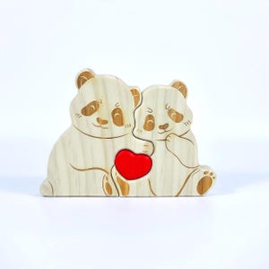 May include: A wooden puzzle featuring two panda bears embracing a red heart. The pandas are light-colored with brown accents, including their ears and facial features. The puzzle is designed to interlock, creating a charming display.