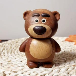 Puede incluir: Figura de oso de madera hecha a mano con vientre marrón claro y nariz y cejas marrones oscuras. El oso tiene una cabeza redonda, orejas pequeñas y está de pie. La figura es de madera y tiene un acabado liso.