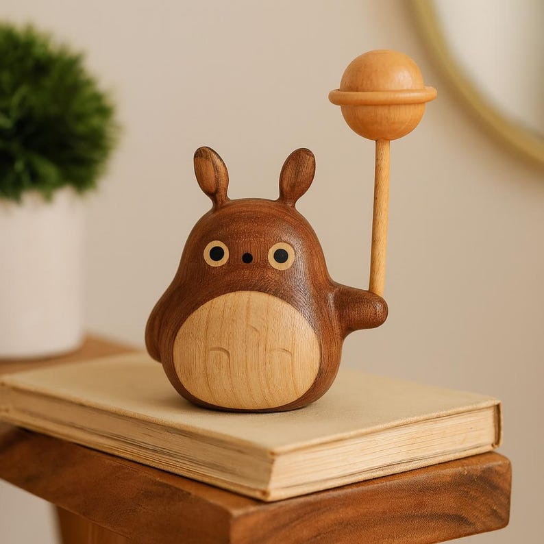 May include: A wooden figurine of a cartoon character with a round body, small ears, and large eyes. The figurine is holding a wooden lollipop. The figure is brown with a lighter wood belly and is sitting on a book.