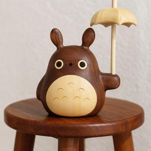 May include: A wooden figurine of a cartoon character with a brown body, large ears, and a light-colored belly. The character holds a small, light-colored wooden umbrella. The figurine is sitting on a small, round wooden stool.
