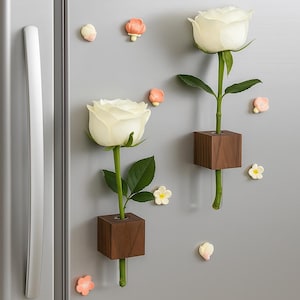 Könnte beinhalten: Nahaufnahme einer Kühlschranktür mit zwei weißen Rosen in kleinen, quadratischen Holzvasen. Mehrere kleine, dekorative Blumenmagnete in Rosa- und Weißtönen sind um die Rosen herum verstreut. Die Kühlschranktür ist grau.