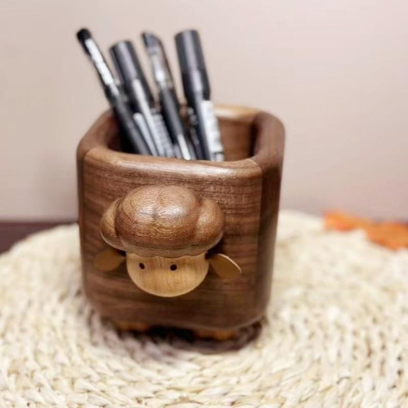 May include: A wooden pen holder with a sheep design. The pen holder is made of dark brown wood and features a carved sheep head on the front. Several black pens are inside the holder. The pen holder sits on a woven mat.