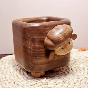 May include: A square wooden container with a carved sheep design. The container is a dark brown color, with a smooth finish. The sheep is light brown with small ears and a round head. The container sits on four small wooden feet.