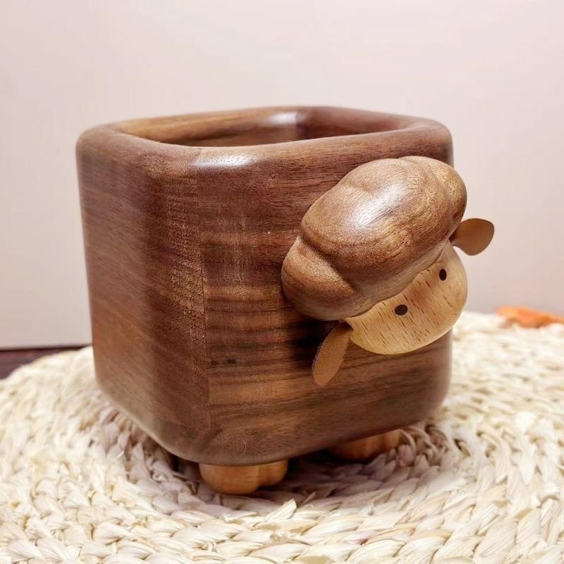 May include: A square wooden container with a carved sheep design. The container is a dark brown color, with a smooth finish. The sheep is light brown with small ears and a round head. The container sits on four small wooden feet.