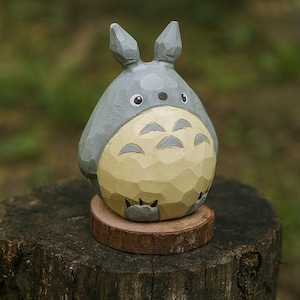 May include: A hand-carved wooden figurine of Totoro, a character from the animated film My Neighbor Totoro. The figure is gray with a cream-colored belly and sits on a small, round wooden base. The figurine is displayed on a tree stump.