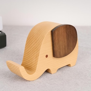 May include: A wooden elephant-shaped phone stand. The stand is made of light-coloured wood with a dark brown ear and a small red dot for an eye. The phone rests on the elephant's trunk, which curves upwards.