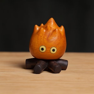 May include: A wooden sculpture of a cartoon fire demon with orange flames and yellow eyes. The figure sits on a dark brown wooden base. The background is a dark color, and the sculpture is on a light brown surface.