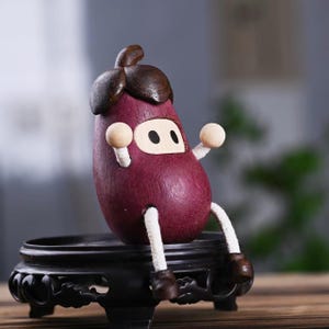 Könnte beinhalten: Eine skurrile Holz-Auberginen-Figur mit einem cartoonartigen Gesicht und Gliedmaßen. Die Aubergine ist tiefviolett mit braunen Akzenten und sitzt auf einem kleinen, verzierten schwarzen Ständer. Arme und Beine bestehen aus weißem Seil und Holzkugeln.