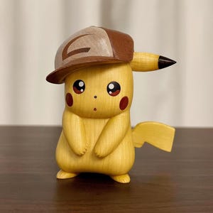 May include: A wooden Pikachu figurine wearing a brown and tan baseball cap. The yellow figure has red cheeks, black eyes, and a black-tipped tail. The figurine is carved with smooth details and stands on a dark surface.