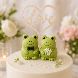 May include: A cake topper featuring two green frog figurines on a white cake. One frog wears a black bow tie, and the other wears a white veil and holds a small bouquet of white flowers. A wooden heart-shaped sign with the word love is centered.